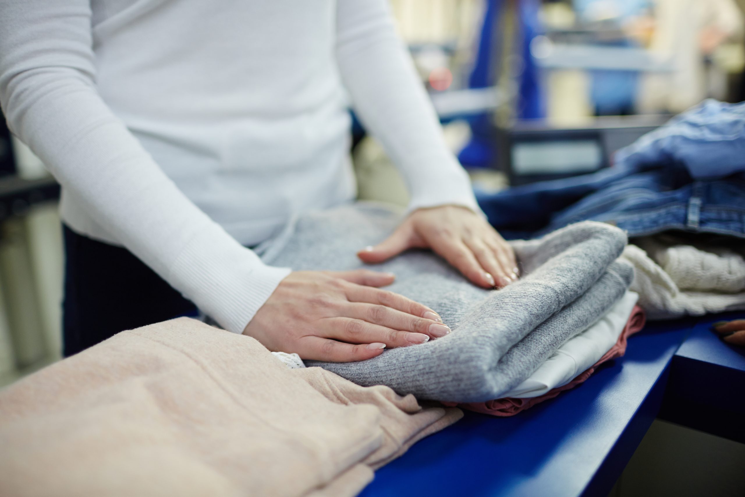 A person in a white long-sleeve shirt neatly folding a stack of clothes on a blue table.